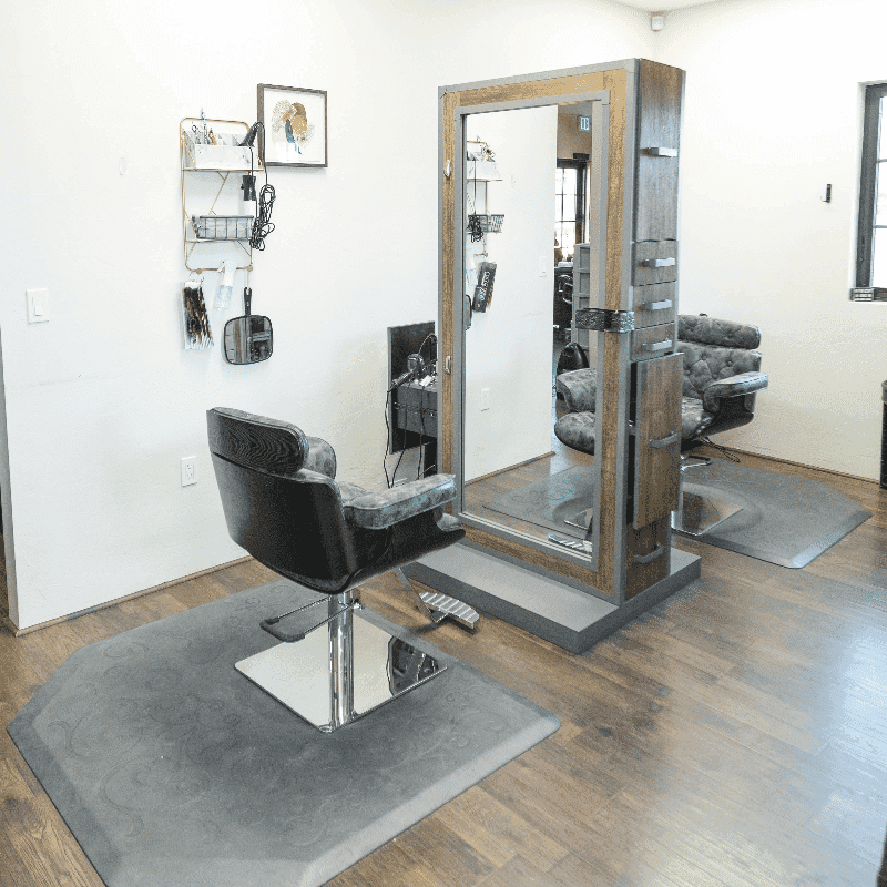 Modern, empty hair salon with a single styling station, mirror, and black chair on wood flooring.